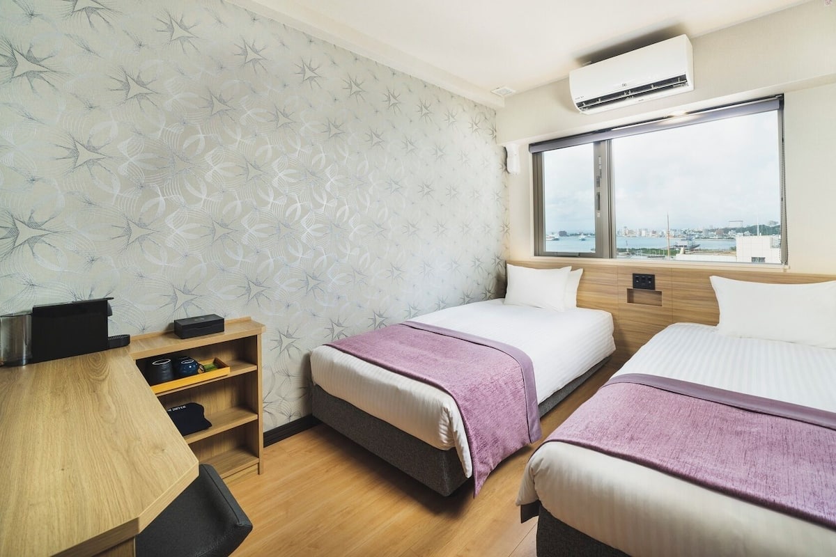 A hotel room with two beds, a desk, a shelf with items, and a window with a city view.