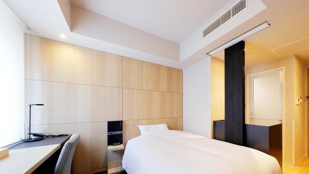 A modern hotel room with a large bed, a desk with a chair, a small table, and a wall-mounted heater. - JR Inn Sapporo N2 (Sapporo)