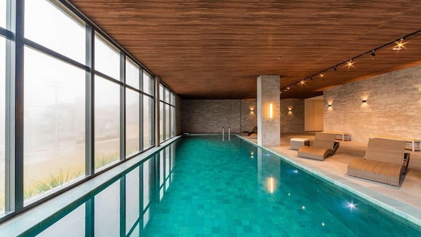 A modern indoor swimming pool with a wooden ceiling, large windows, and lounge area with seating. - Laghetto Viverone Canela (Canela)