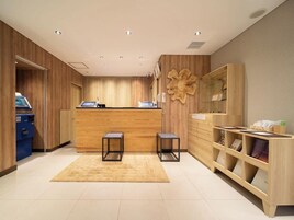 Una zona de recepción con un mostrador de madera, dos taburetes negros, un expositor de vidrio y una máquina azul, probablemente un quiosco de autoservicio.