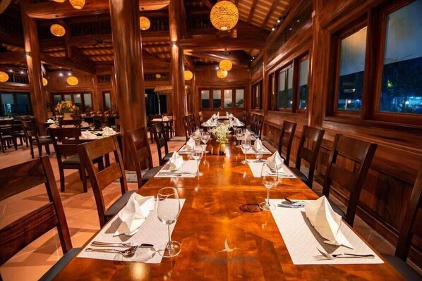 Una larga mesa de comedor de madera, cubierta con manteles y servilletas blancas, y cubiertos, rodeada de sillas de madera en un restaurante con grandes ventanales y lámparas colgantes.