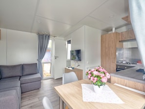 Ein modernes, kompaktes Wohnzimmer mit Sofa, Esstisch mit Blumenvase, Küchenzeile und Fernseher.