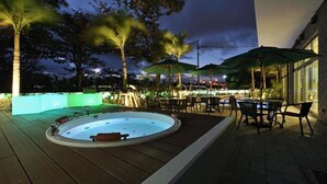 Un espace extérieur aménagé avec un jacuzzi, des sièges et des parasols, entouré d’arbres et d’un bâtiment.