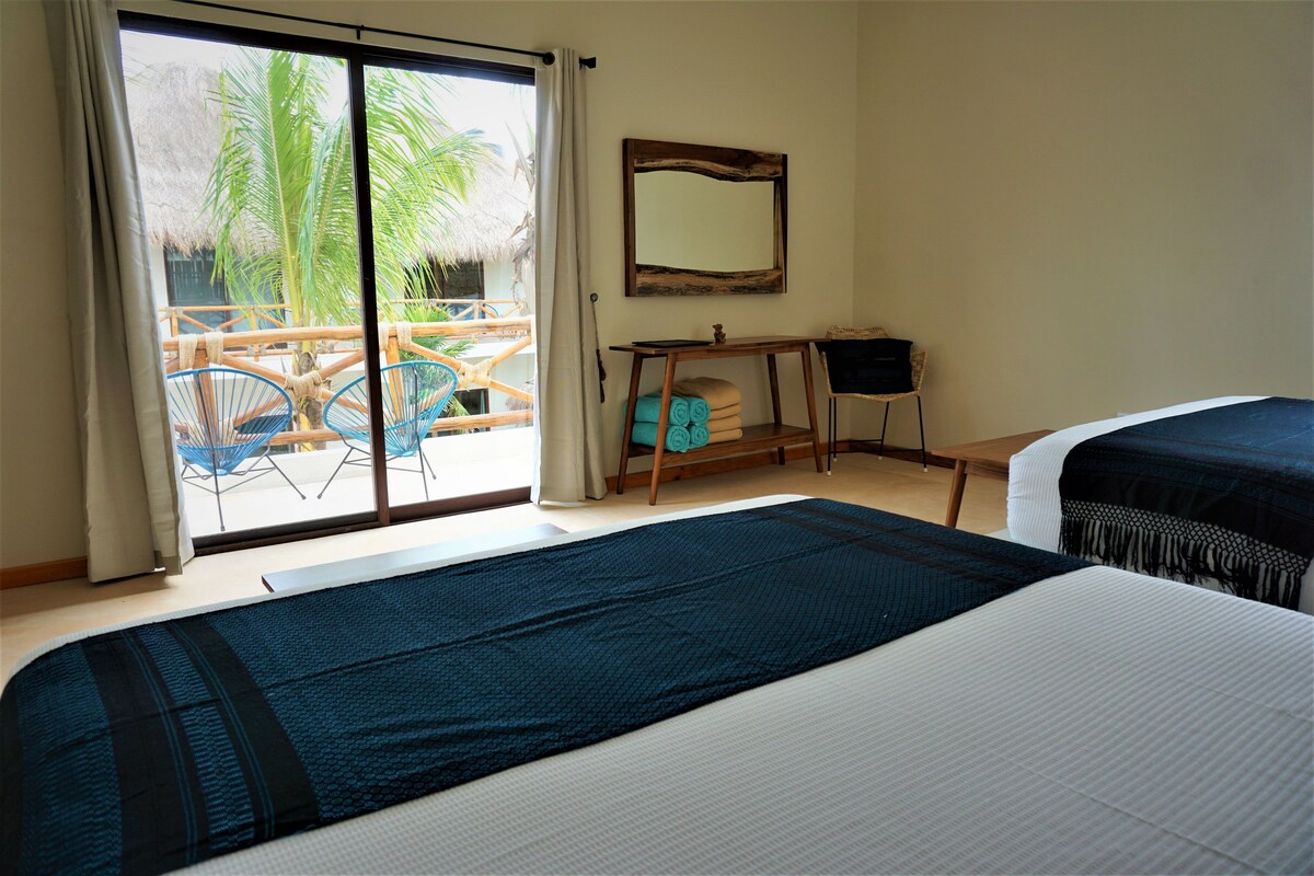 A hotel room with a bed, a desk, and a view of a palm tree and thatched roof structure outside.