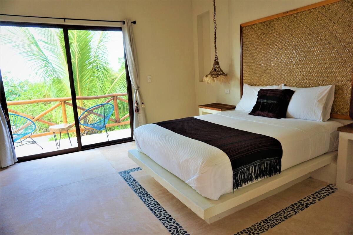 A bedroom with a large bed, a nightstand, a hanging lamp, and a view of palm trees through a sliding glass door.