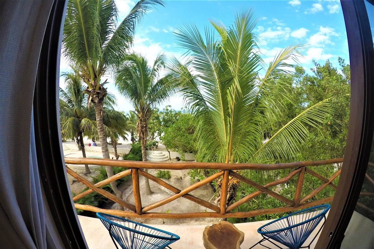 A balcony with a wooden railing, two blue wire chairs, and a view of palm trees and greenery.