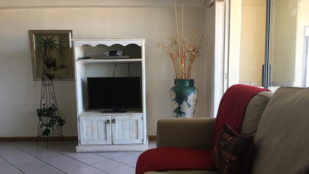 A living room with a beige sofa, a white entertainment unit with a flat-screen TV, a decorative vase, and a tall plant. - Seashells Holiday Self Catering Apartments (Jeffreys Bay)