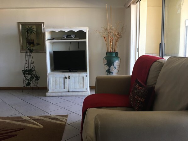 A living room with a beige sofa, a white entertainment unit with a flat-screen TV, a decorative vase, and a tall plant. - Seashells Holiday Self Catering Apartments (Jeffreys Bay)