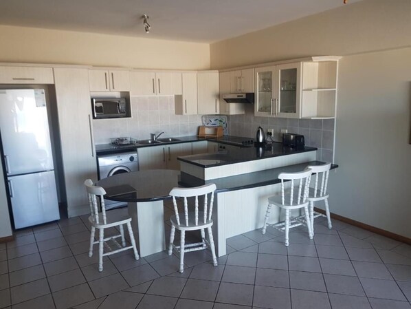 A compact kitchen with a central island, white bar stools, a built-in microwave, and a refrigerator. - Seashells Holiday Self Catering Apartments (Jeffreys Bay)