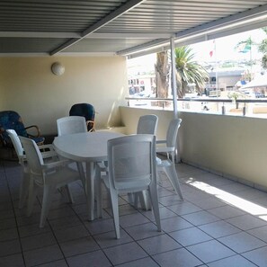 A covered patio with a round table and six chairs, a sofa, and a view of the street. - Seashells Holiday Self Catering Apartments (Jeffreys Bay)