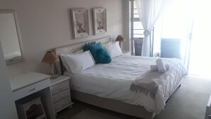A bedroom with a bed, bedside table, lamp, and a window with curtains. - Seashells Holiday Self Catering Apartments (Jeffreys Bay)