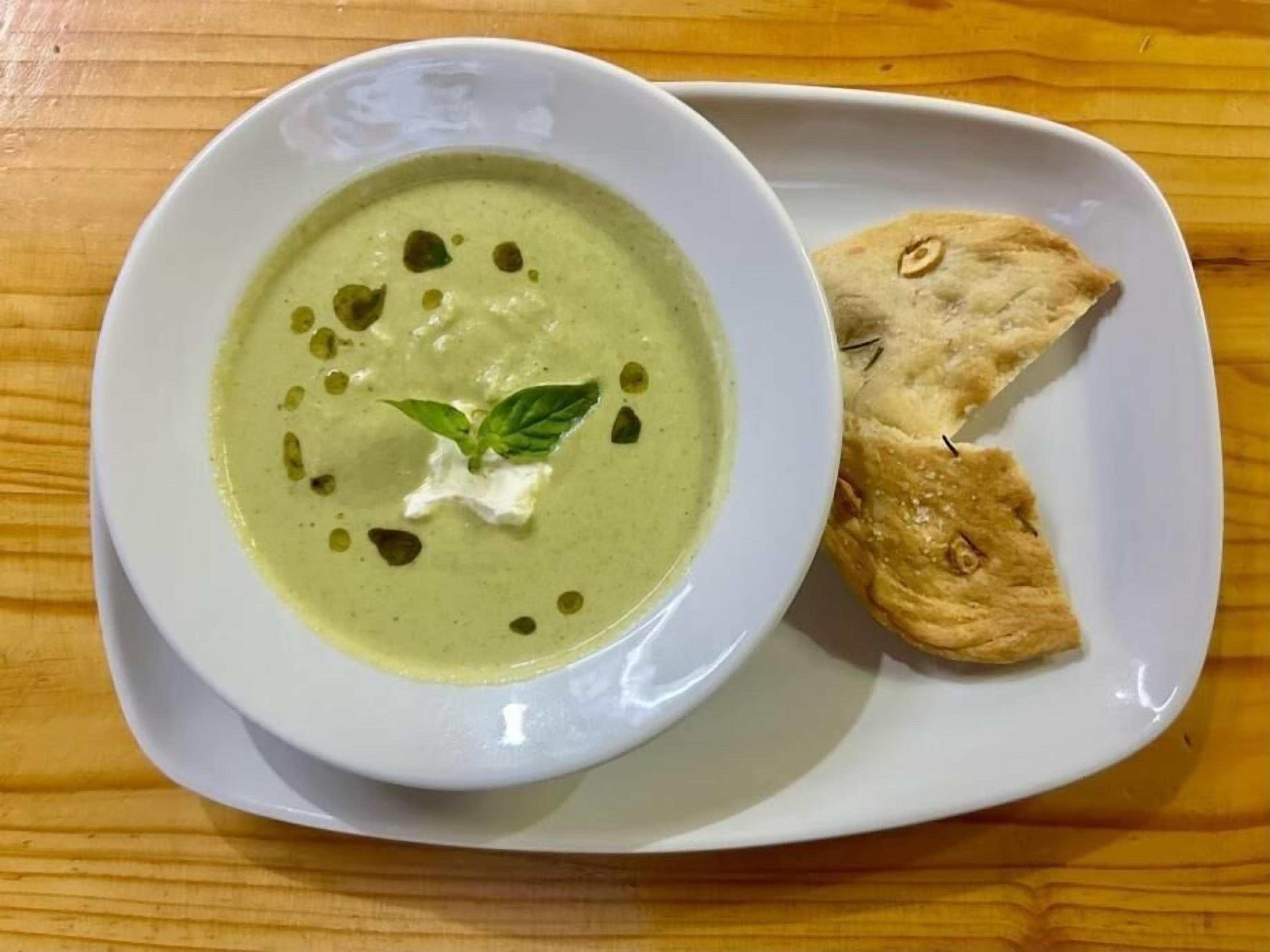 A bowl of green soup with a dollop of cream and a basil leaf on a white plate, accompanied by two pieces of bread.
