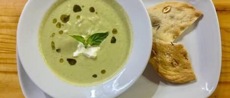 A bowl of green soup with a dollop of cream and a basil leaf on a white plate, accompanied by two pieces of bread.