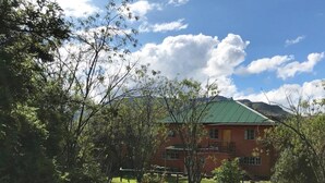 Un bâtiment rouge surmonté d’un toit vert, entouré d’arbres et d’herbe, avec des montagnes en arrière-plan.