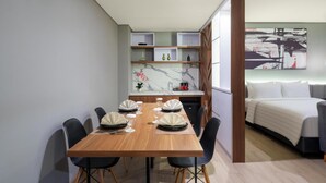 A modern hotel room with a dining area, a bed with a white comforter, and a marble backsplash in the kitchen.