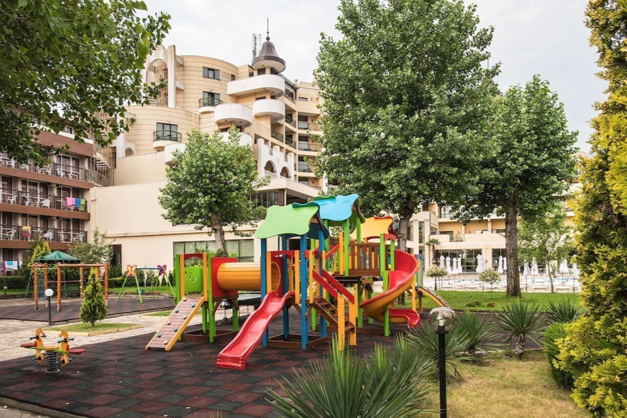 Ein Spielplatz mit Rutschen und Klettergerüsten, umgeben von Bäumen und einem Gebäude mit Balkonen im Hintergrund.