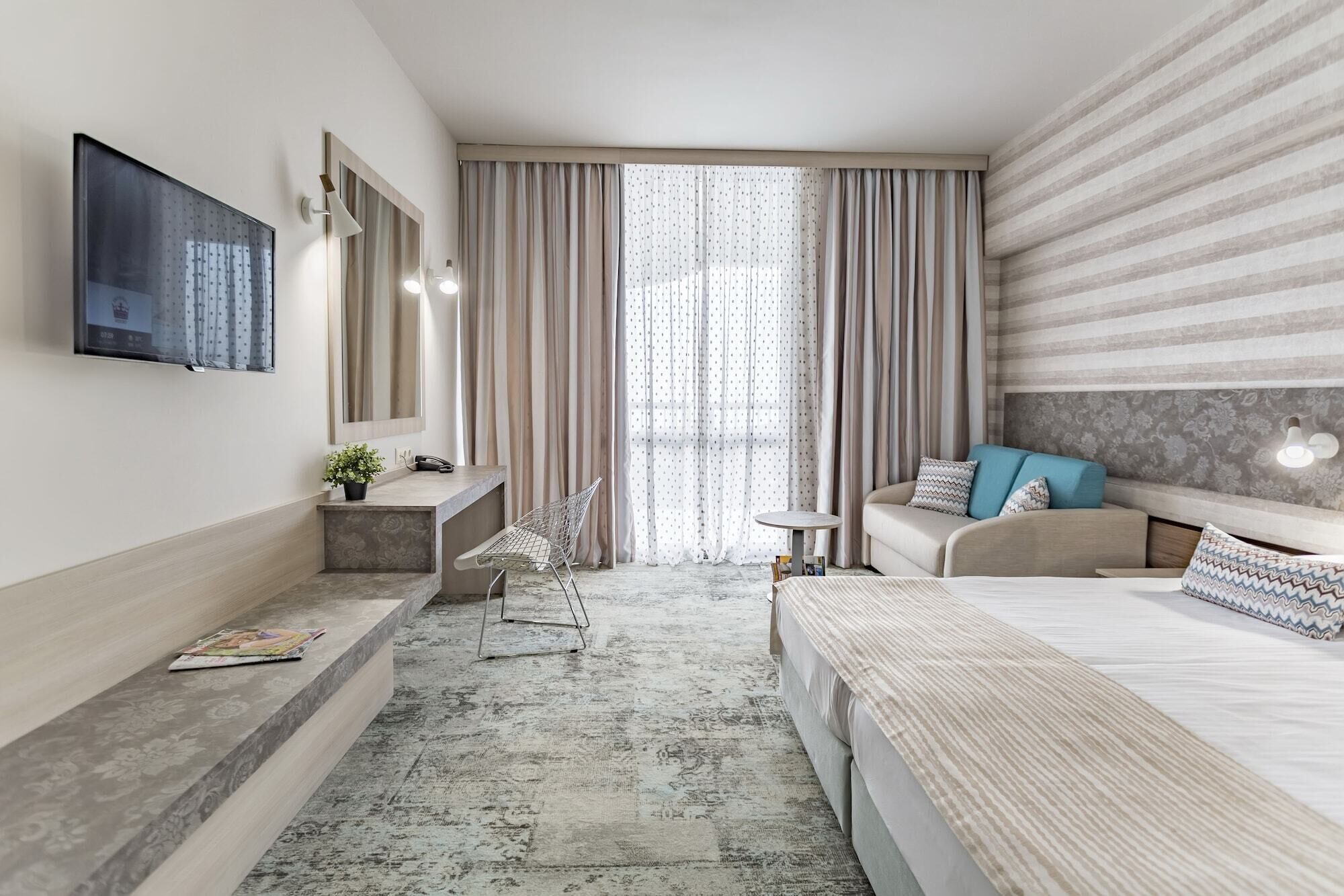 A modern hotel room with a large bed, a sofa, a desk with a chair, a television, and a window with curtains.