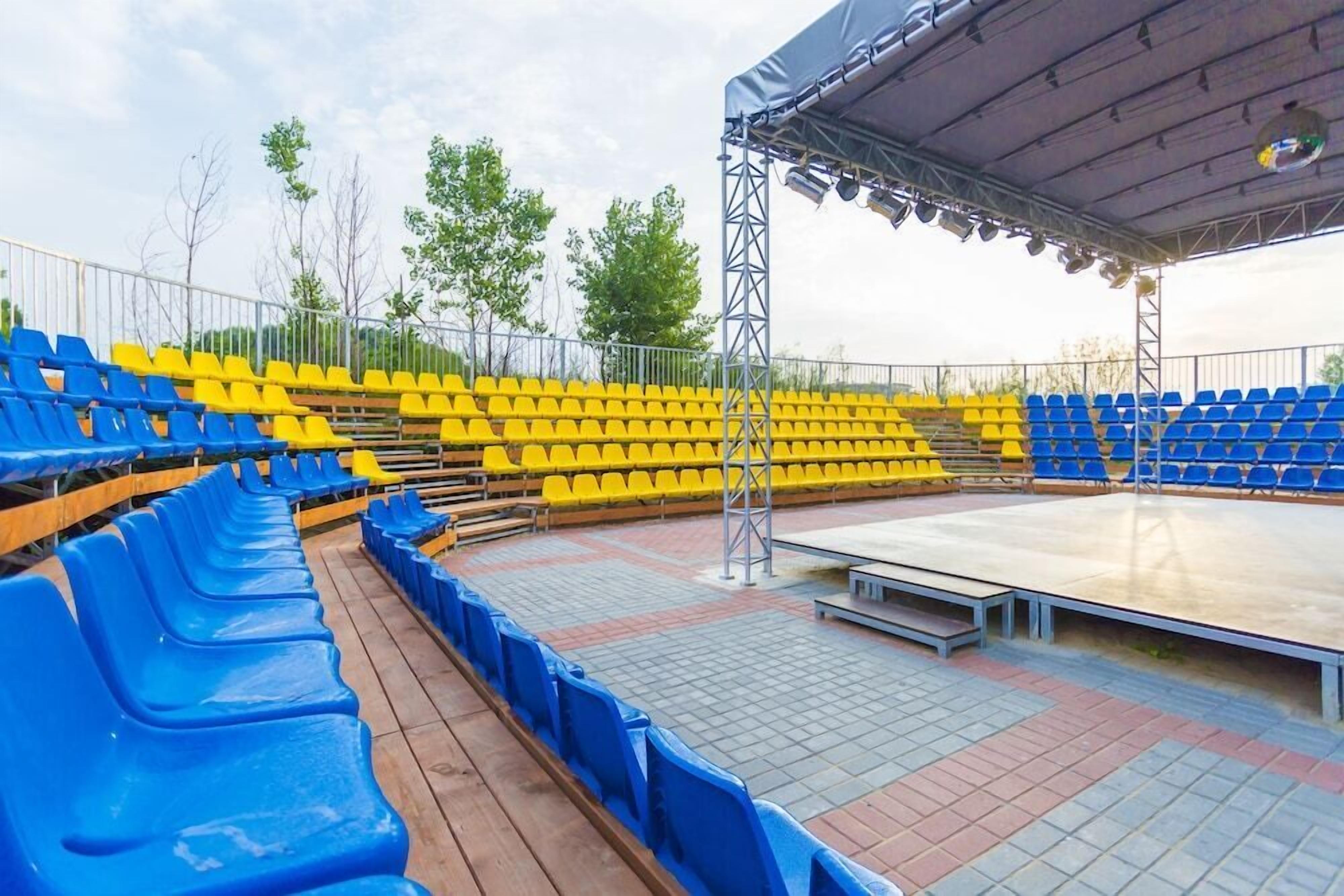 An outdoor amphitheater with tiered seating in blue and yellow, a stage, and a metal railing.
