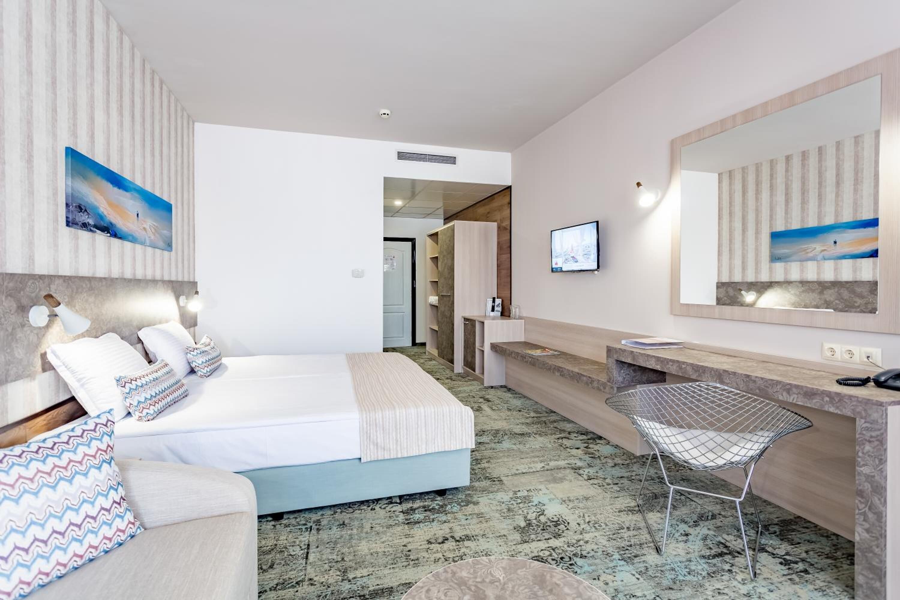 A hotel room with a bed, a desk, a chair, a TV, and artwork on the walls.