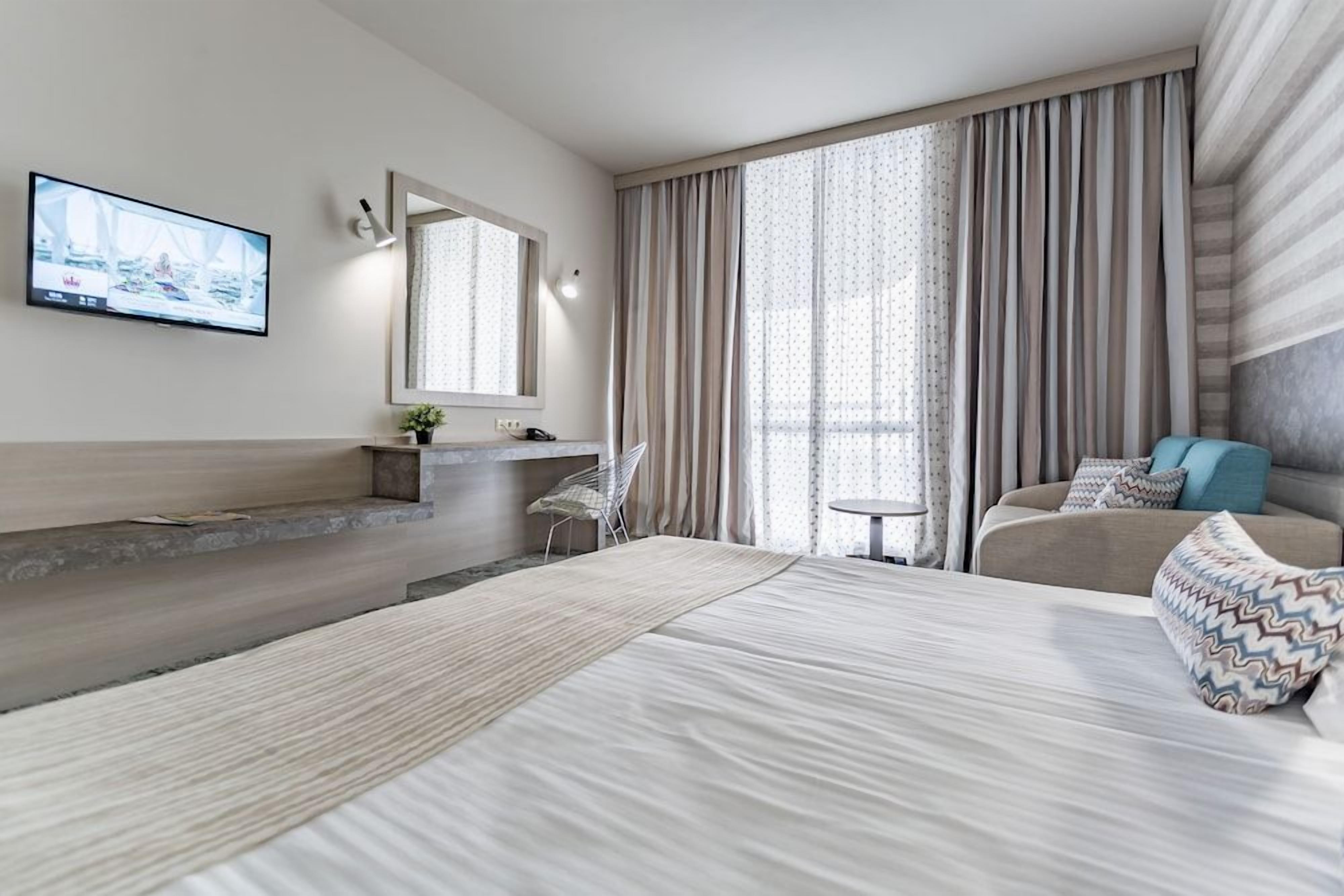 Habitación de hotel con una cama grande, un televisor montado en la pared, una mesita, un sofá y cortinas.