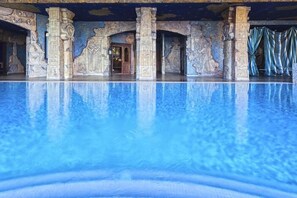 Een groot overdekt zwembad met helderblauw water, omringd door stenen zuilen met oude Egyptische gravures, en een deur naar een andere ruimte.