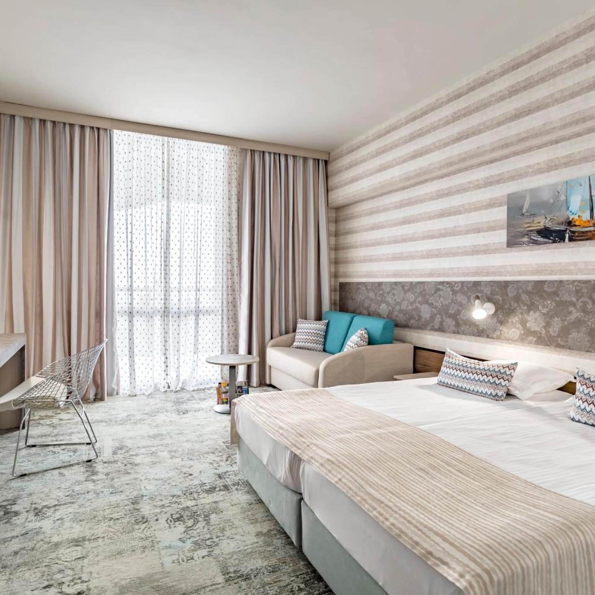 Habitación de hotel moderna con una cama grande, una zona de estar con sofá y sillones, una mesita y un ventanal con cortinas.