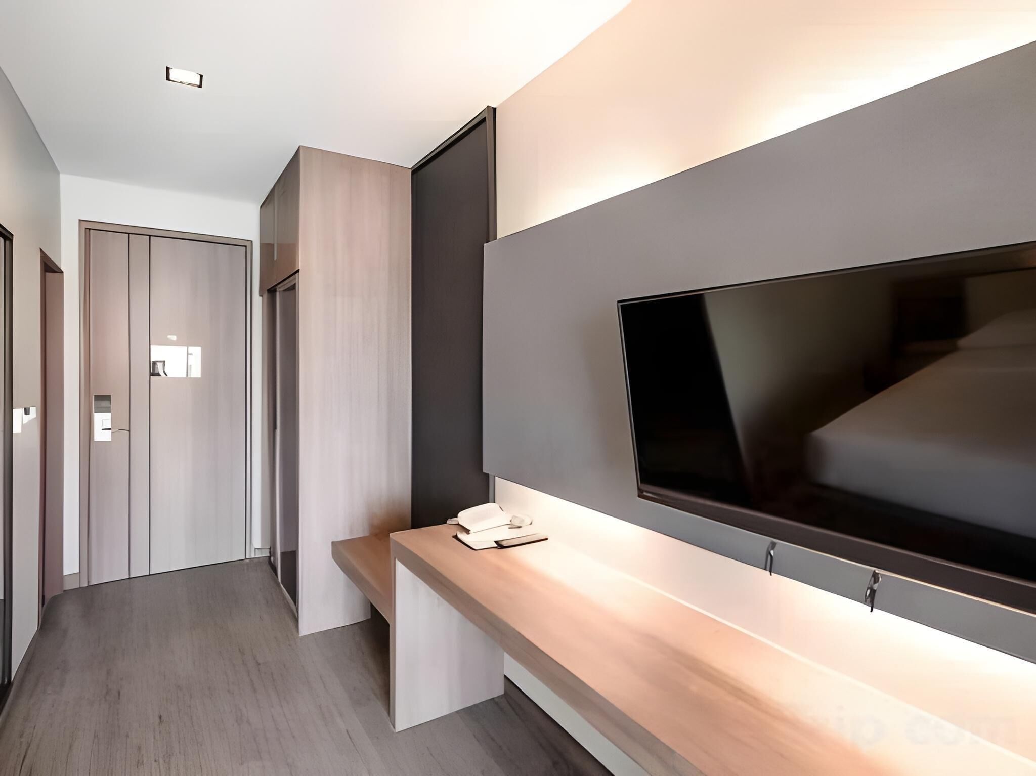 A modern hotel room with a large flat-screen TV mounted on a wall, a wooden console table, and a door with a light switch.