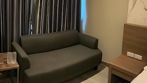 A modern hotel room with a dark green sofa, a wooden side table, and a small bedside table with a lamp. - The Chess Hotel (Rayong)