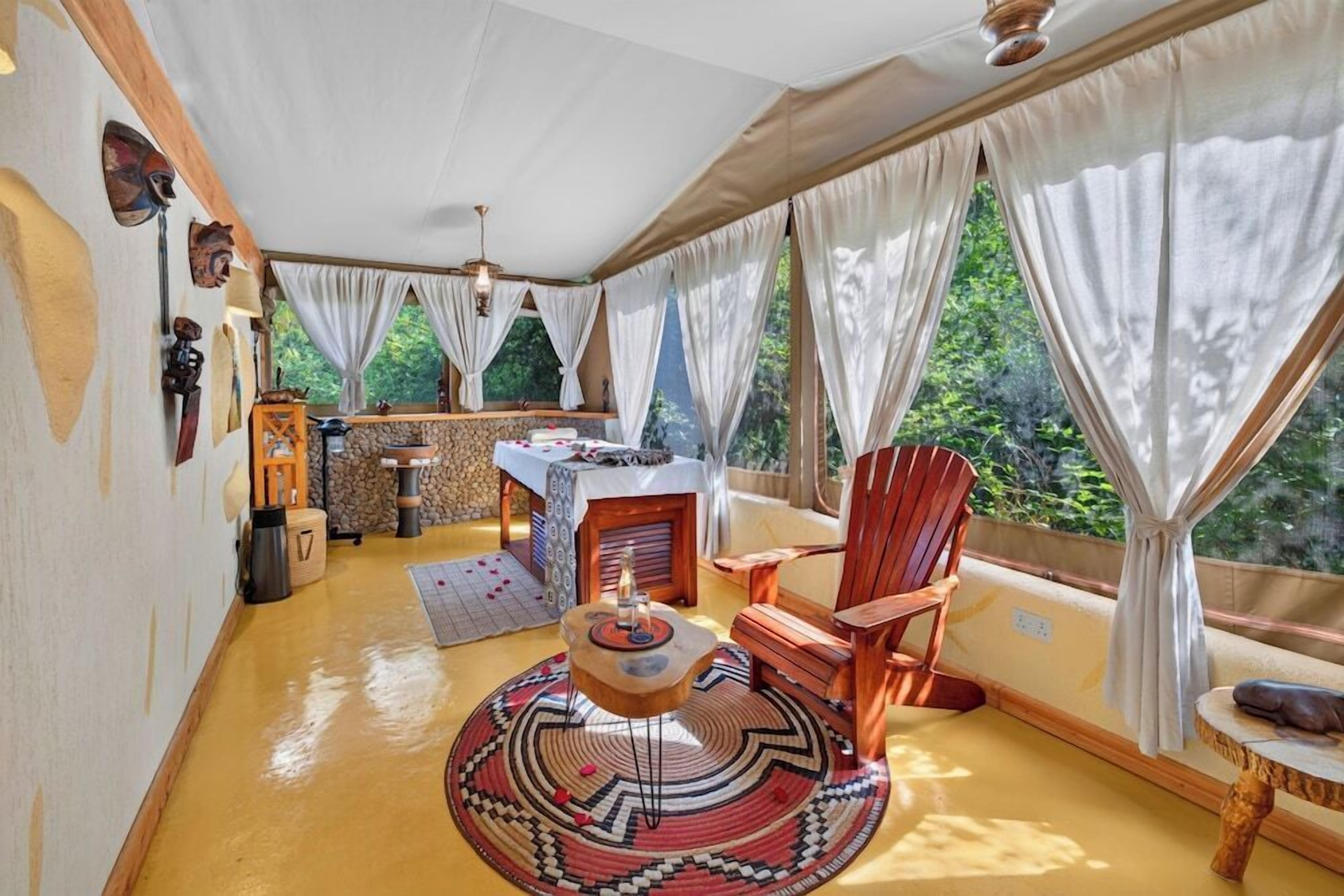A cozy room with a wooden floor, a round table with a rug, a wooden chair, and a sofa. There are curtains on the windows, and a view of greenery outside.