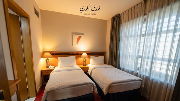 A hotel room with two beds, a wooden headboard, a nightstand with a lamp, and a window with curtains. - Kindi Suites Hotel (Amman)