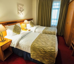 A hotel room with two beds, a nightstand, a lamp, a wall art piece, and a window with curtains. - Kindi Suites Hotel (Amman)