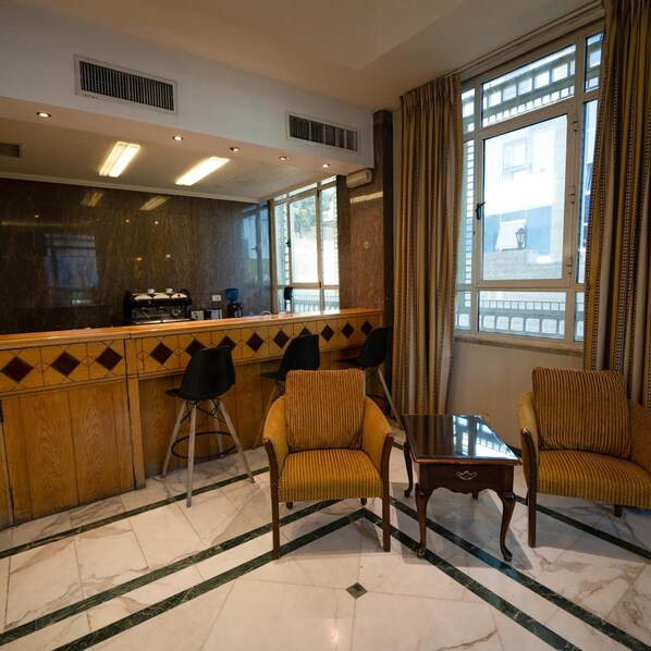 A hotel lobby with a reception desk, two chairs, a small table, and large windows with curtains. - Kindi Suites Hotel (Amman)