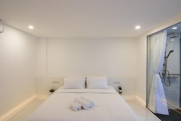 Moderni hotellihuone, jossa on suuri sänky, valkoiset vuodevaatteet, lasiseinäinen suihkutila ja pieni ruukkukasvi.