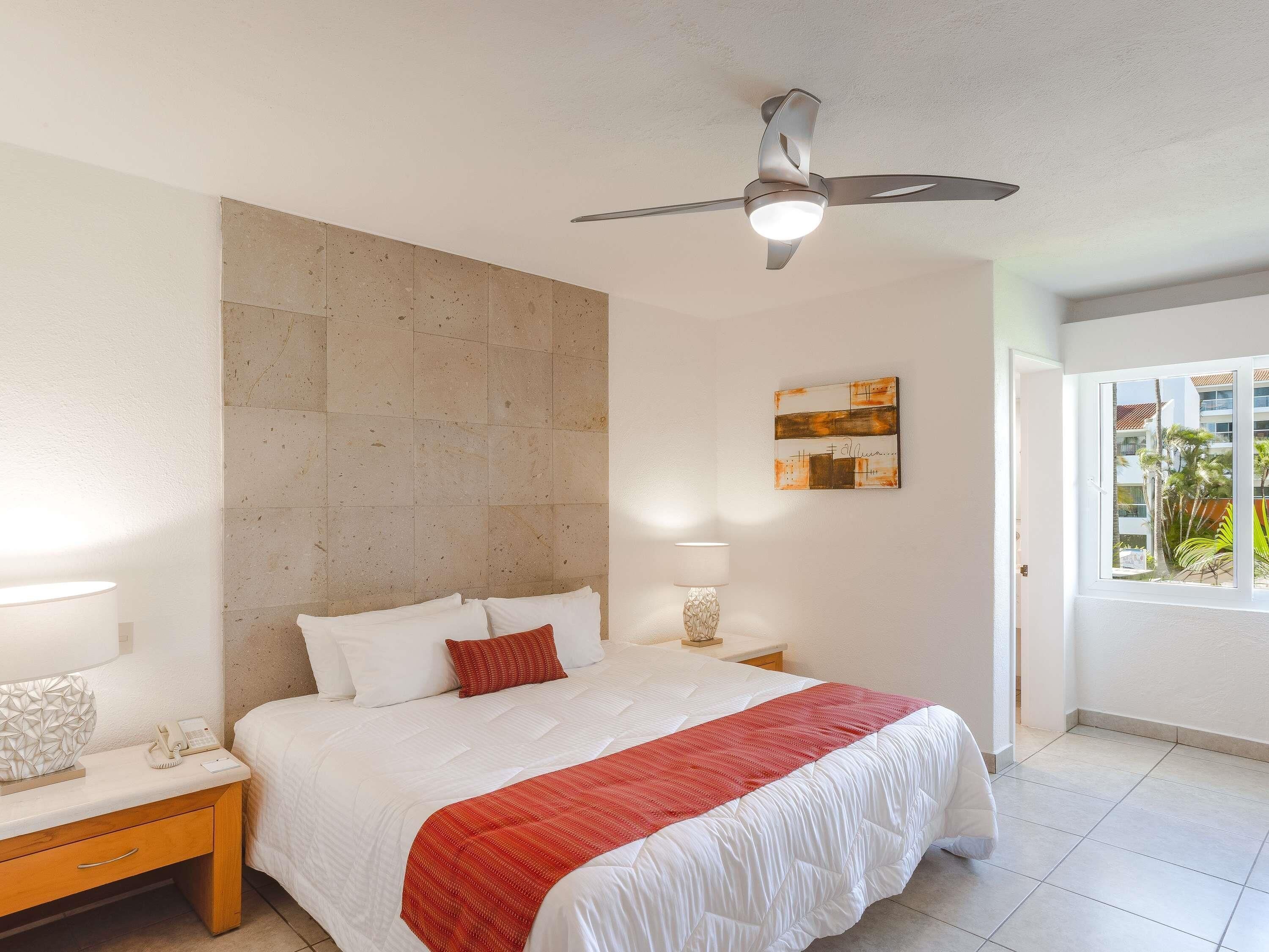 Habitaci ón de hotel con cama, lámparas de noche, ventilador de techo, ventana con vistas y una obra de arte colgada en la pared.