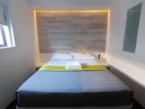 A hotel room with a bed, a headboard, a window, and a wall-mounted light fixture. - Check Inn Hotel (Cape Town)
