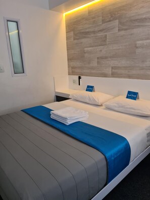 A hotel room with a bed, a nightstand, a wall-mounted light, and a window. - Check Inn Hotel (Cape Town)