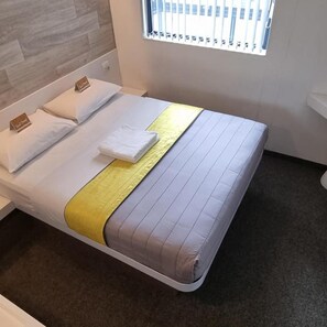 A neatly made bed with pillows and a folded towel, a window with blinds, and a wooden wall. - Check Inn Hotel (Cape Town)