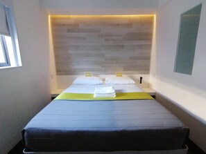A hotel room with a bed, a headboard, a window, and a wall-mounted light fixture. - Check Inn Hotel (Cape Town)