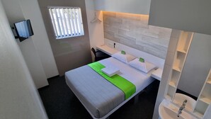 A compact hotel room with a bed, a desk, a chair, a small window, and a bathroom with a sink and mirror. - Check Inn Hotel (Cape Town)