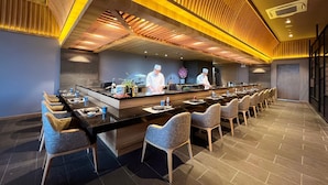 A modern restaurant with a long counter, multiple chairs, and two chefs preparing food. - Da Nang Mikazuki Japanese Resorts and Spa (Da Nang)