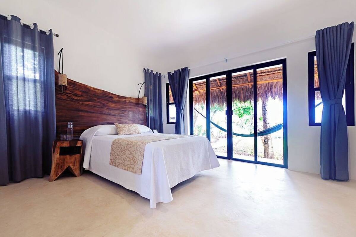 A spacious bedroom with a large bed, a wooden headboard, and a view of a thatched roof structure outside.