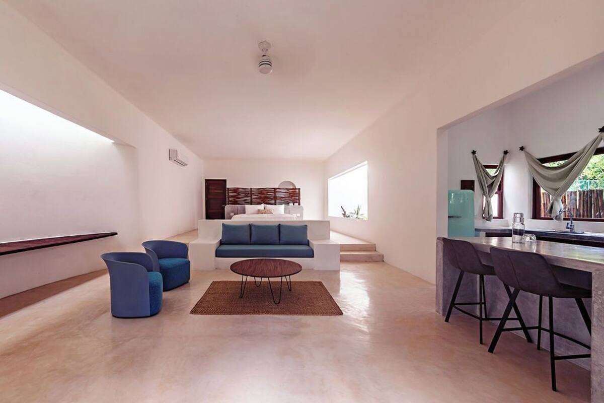 A modern living room with a blue sofa, two blue armchairs, a coffee table, and a kitchen in the background.