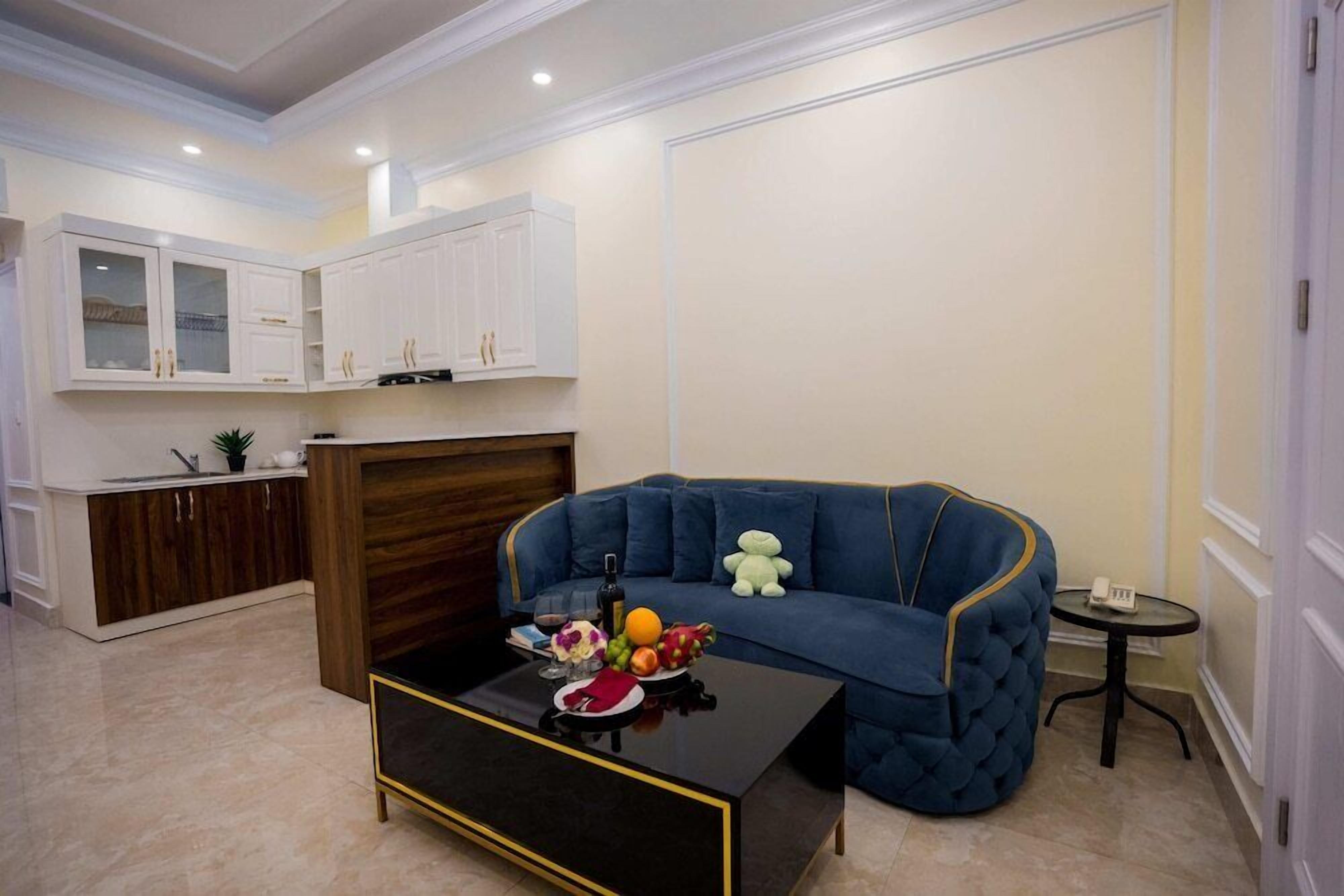 A modern living room with a blue sofa, a coffee table with fruit, and a kitchen area with white cabinets.