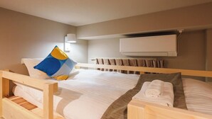 Un lit simple avec un cadre en bois, une literie blanche, un oreiller bleu et jaune, une serviette enroulée et un climatiseur fixé au mur.
