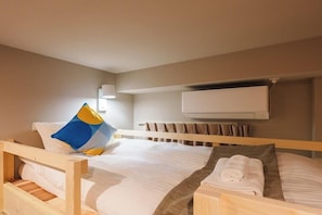 A single bed with a wooden frame, white bedding, a blue and yellow pillow, a rolled towel, and a wall-mounted air conditioner.