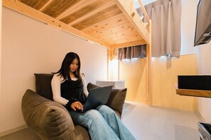Una persona sentada en un sofá usando una computadora portátil en una habitación con vigas de madera en el techo, un televisor de pantalla plana montado en la pared y una ventana con cortinas.