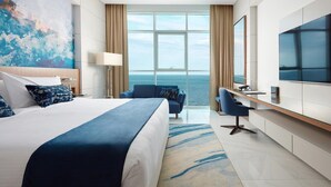 A modern hotel room with a large bed, a desk, a chair, a TV, and a view of the ocean. - Royal M Resort Al Aqah Beach Resort (Al Aqah)