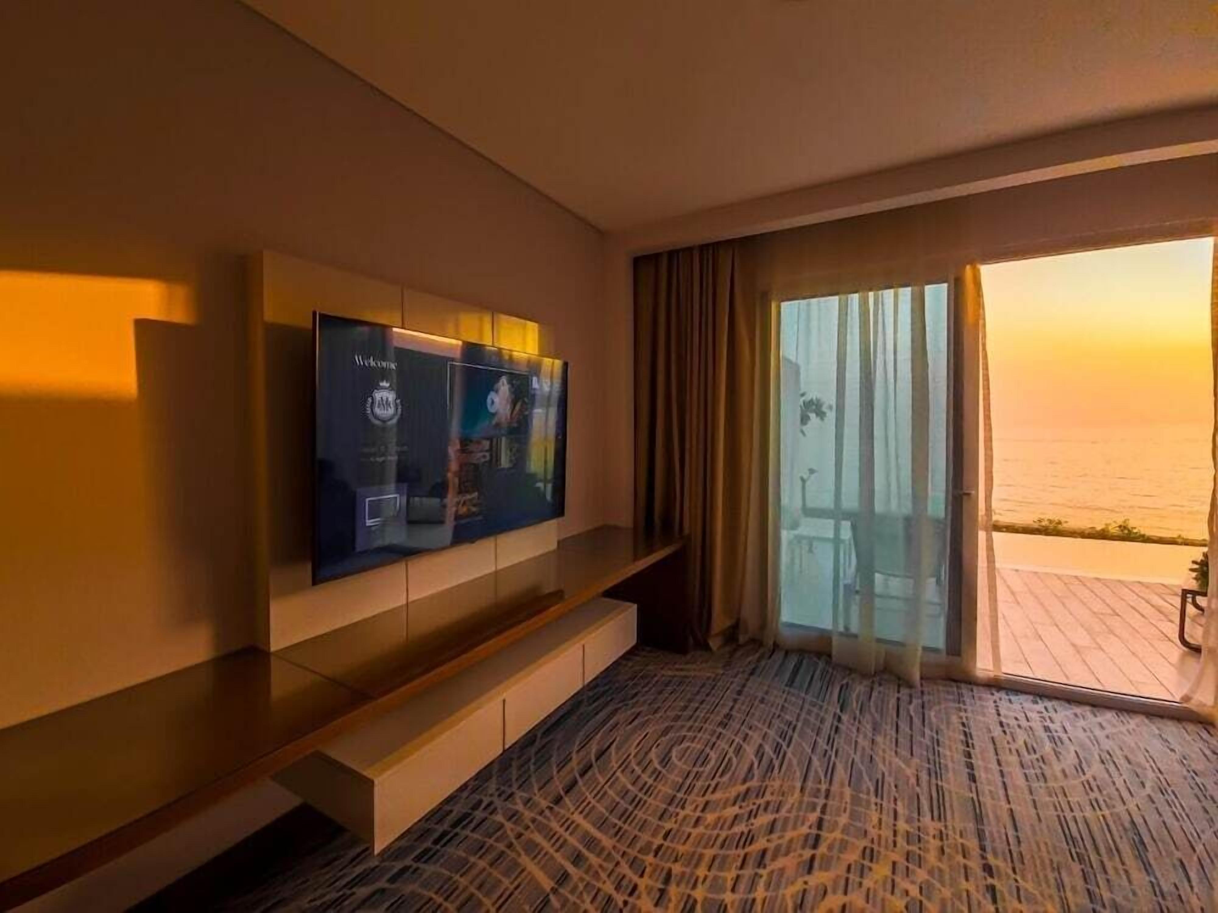 A hotel room with a large TV on the wall, a balcony with a view of the sea, and a patterned carpet.