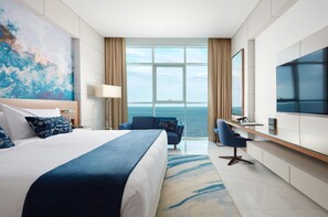 A modern hotel room with a large bed, a desk, a chair, a TV, and a view of the sea.