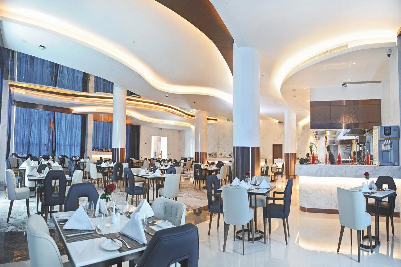 A spacious hotel dining area with multiple tables and chairs, a central bar, and a modern kitchen visible in the background.